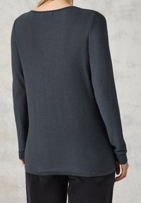 Personne portant un haut texturé à manches longues gris foncé et un pantalon en velours côtelé noir, debout devant un fond clair et tacheté.