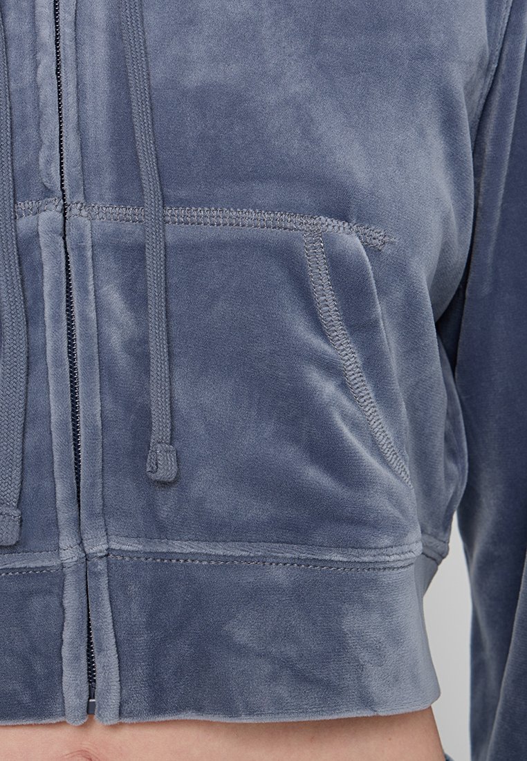 Verkorte, gevoerde jas van zacht blauwe stof. Voorzien van een ritssluiting aan de voorkant, een trekkoord en twee voorzakken met zichtbare stiksels.