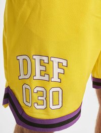 Geltonos tinklinės šortai su balta ir purpurine tekstu "DEF 030" apatinėje kairėje, su purpurinėmis ir juodomis dryžuotomis juostelėmis palei kraštą.