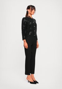 Mujer con coleta oscura lleva un cárdigan de lentejuelas negras sobre una blusa de estampado de leopardo, pantalones negros recortados y tacones de kitten black, posando de lado.