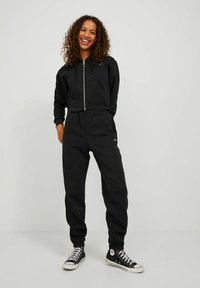 Black cropped hoodie with a front zipper and pocket, paired with black joggers featuring an elastic waistband and cuffed ankles, worn with black sneakers.
