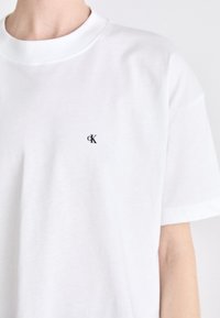 Bijela pamučna majica s okruglim izrezom i kratkim rukavima. Mali crni "ck" logo je vezan na lijevoj strani prsnog dijela.