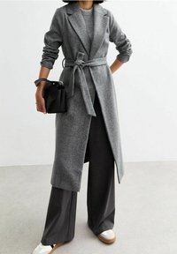 Abrigo largo de espiga gris con cintura ajustada, combinado con pantalones gris de pierna ancha y zapatillas blancas. Un bolso negro completa el conjunto.
