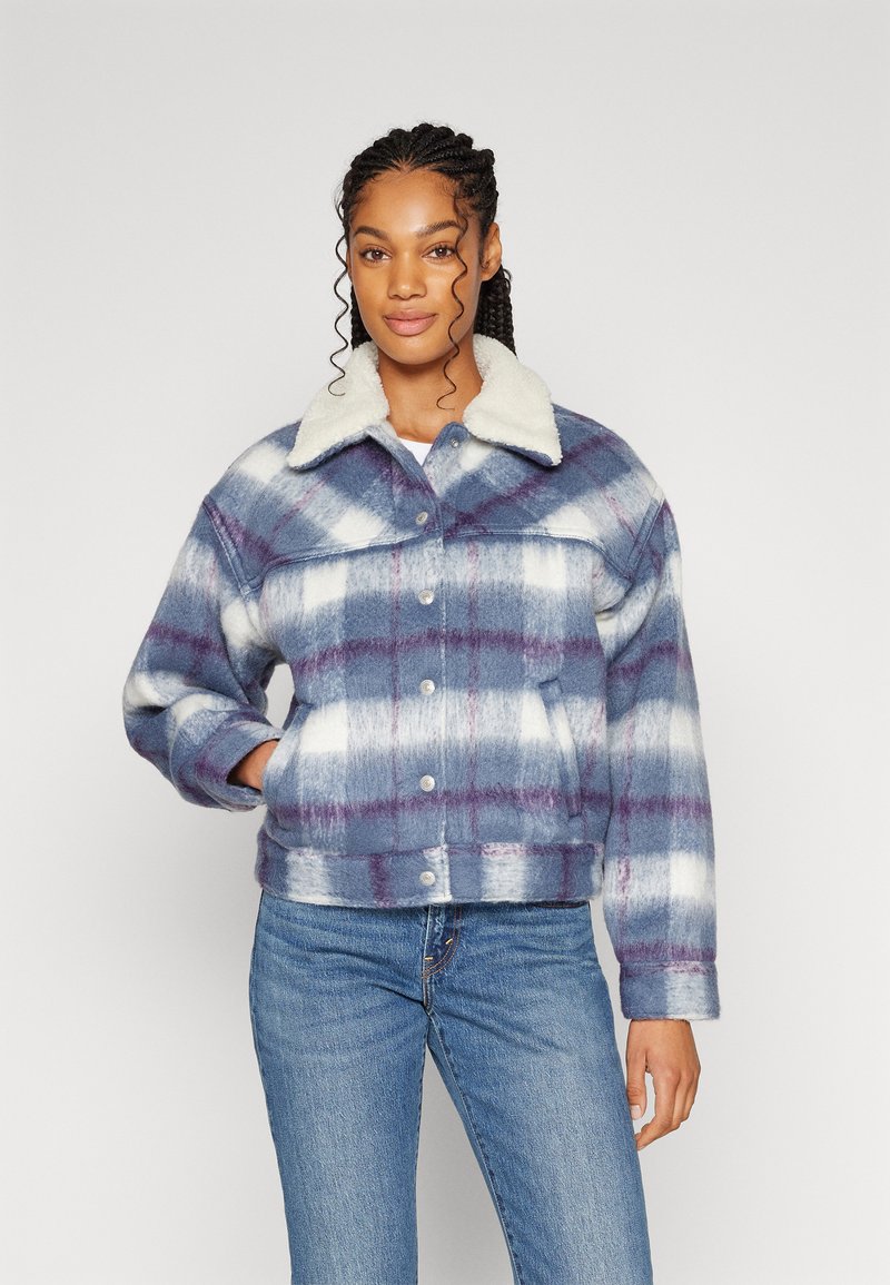 Femme portant une veste en plaid bleu et blanc avec un col en sherpa et un jean bleu, se tenant avec une main dans la poche devant un fond uni.