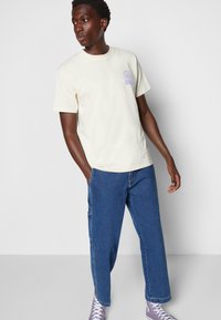 Cream-colored cotton t-shirt featuring a front purple graphic design, paired with blue denim pants and gray sneakers.