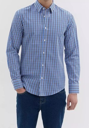 Hombre con una camisa de manga larga a cuadros azules y blancos y vaqueros azul oscuro, de pie con una mano en el bolsillo.