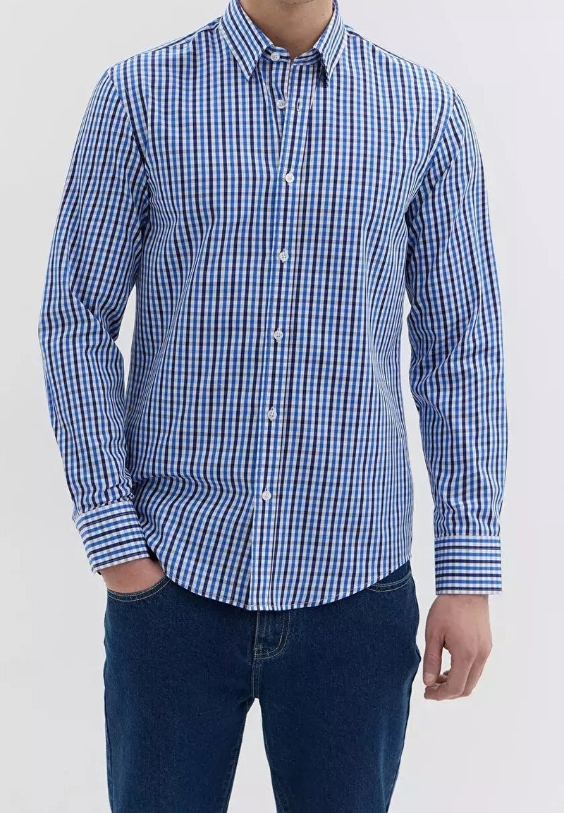 Homme portant une chemise à manches longues à carreaux bleus et blancs et un jean bleu foncé, debout avec une main dans la poche.