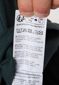 Etiket fra et mørkegrønt beklædningsstykke, der viser plejeinstruktioner og stofsammensætning: 95% bomuld, 5% elastan; tekst på flere sprog.