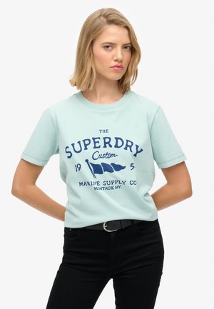 Jasnoniebieska bawełniana koszulka z krótkim rękawem, z ciemnoniebieskim tekstem graficznym i flagami. W połączeniu z czarnymi spodniami i paskiem.