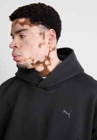 Sort hættetrøje lavet af blødt stof med en front kangaroo-lomme, prydet med et diskret mørkt logo på brystet og en afslappet, oversized pasform.