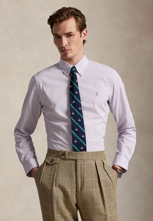 Muž nosí svetlofialovú pruhovanú košeľu na gombíky, námornícku modrú kravatu so zelenými pruhmi a motívmi a hnedé kockované nohavice s vysokým pásom, ruky v vreckách.
