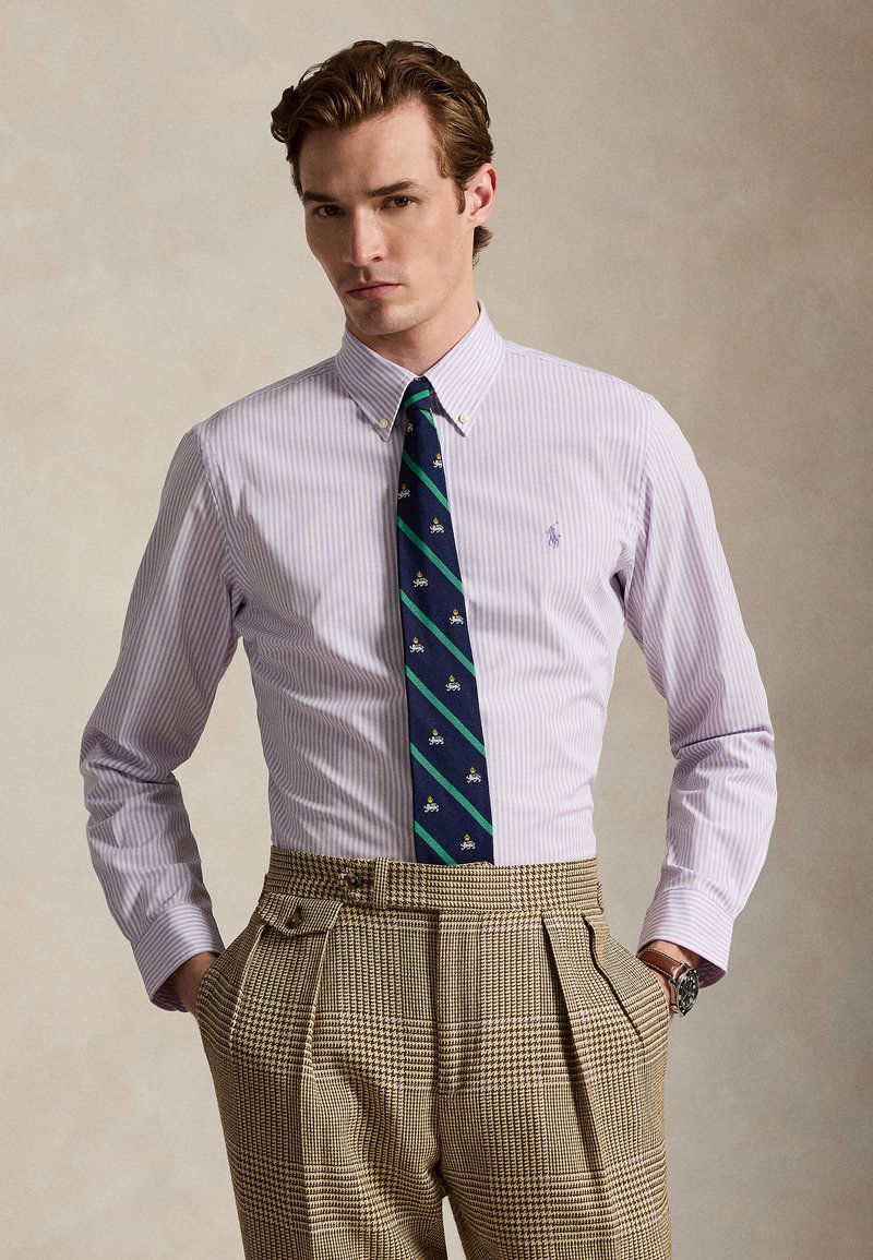 Muž nosí svetlofialovú pruhovanú košeľu na gombíky, námornícku modrú kravatu so zelenými pruhmi a motívmi a hnedé kockované nohavice s vysokým pásom, ruky v vreckách.