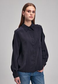 Ciemnoniebieska koszula z długim rękawem, z fakturowanej tkaniny, z jedną kieszonką. Posiada kołnierz typu point oraz ozdobne guziki na ramionach.