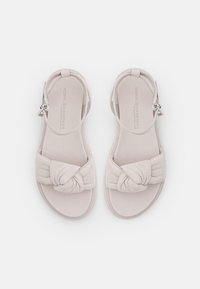 Světlé béžové sandály s měkkým textilním svrškem, se svázaným designem přes přední část a plochou podrážkou. Nastavitelný pásek kolem kotníku pro lepší upevnění.