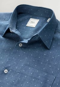 Chemise en chambray bleu avec une coupe régulière, présentant un col boutonné, un petit motif de pois blancs et une poche poitrine. Étiquette de la marque Next visible.