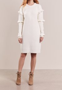 Biała dzianinowa sukienka z prążkowanej tkaniny z falbanami na rękawach, okrągłym dekoltem i długości poniżej kolan. Noszona z brązowymi botkami za kostkę na sznurówki.