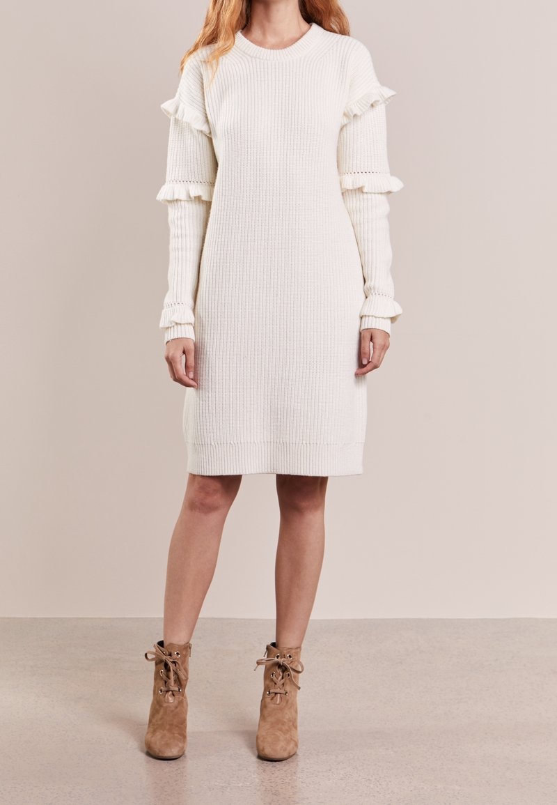 Biała dzianinowa sukienka z prążkowanej tkaniny z falbanami na rękawach, okrągłym dekoltem i długości poniżej kolan. Noszona z brązowymi botkami za kostkę na sznurówki.
