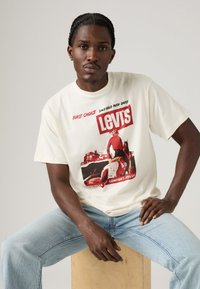 Man met gevlochten haar, gekleed in een romige Levi's graphic t-shirt en lichtblauwe jeans, zittend op een houten blok tegen een effen achtergrond.