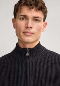 Donkerblauwe geribbelde trui met een hoge kraag en een volledige ritssluiting. Geknoopte stof met zichtbaar patroon en metalen ritssluiting.