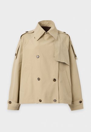 Femme grande aux longs cheveux blonds porte une veste beige à double boutonnage, une chemise bleu clair, un jean large bleu et des baskets rouges, debout sur fond blanc.