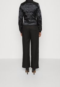 Black quilted jacket with a fitted waist, featuring an embossed logo on the back. Paired with wide-leg black trousers and high-heeled shoes.