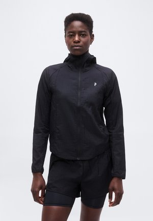 Άτομο που φοράει μαύρο αθλητικό μπουφάν με μακριά μανίκια και φερμουάρ και αντίστοιχα σορτς, στέκεται μπροστά σε απλό φωτεινό φόντο.