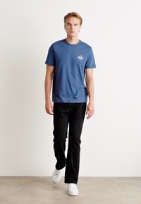 T-shirt blu a maniche corte con una piccola grafica sul petto, abbinato a jeans neri e sneakers bianche, su uno sfondo neutro.