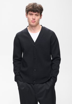 Young man with short brown hair wearing a black buttoned cardigan over a white shirt and black pants, standing with hands in pockets.