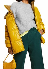 Een gele pufferjack, een grijze gebreide trui, groene broek en een kleine gele handtas. De jas heeft een glanzende afwerking en zichtbare stiksels.