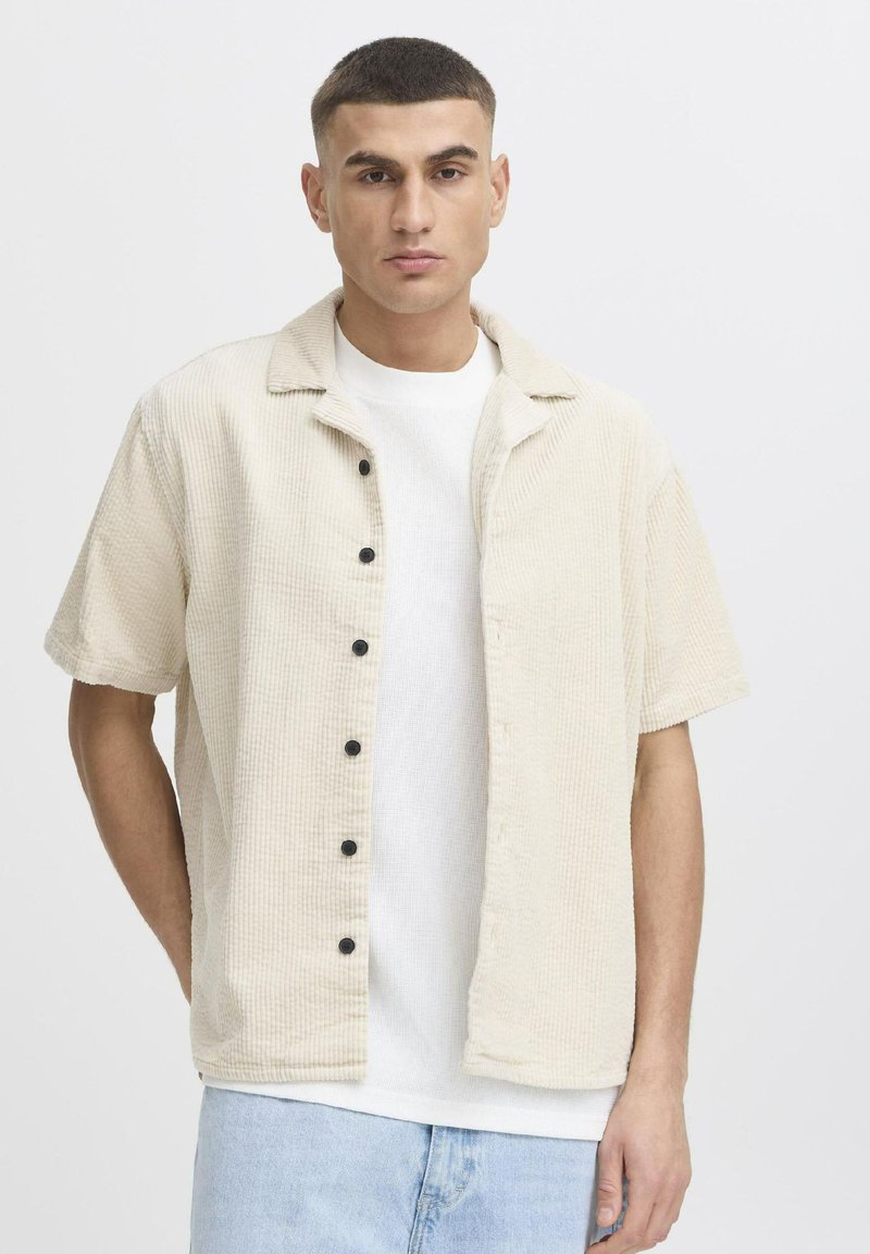 Cream corduroy shirt jacket with a relaxed fit, short sleeves, black buttons, and a flat collar, layered over a white t-shirt.