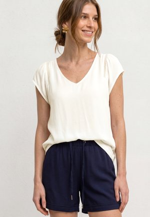 Femme aux cheveux châtain clair noués en chignon, portant des boucles d'oreilles en forme de fleurs, un chemisier blanc à manches courtes et un short bleu marine à cordon, souriante et regardant vers la droite.