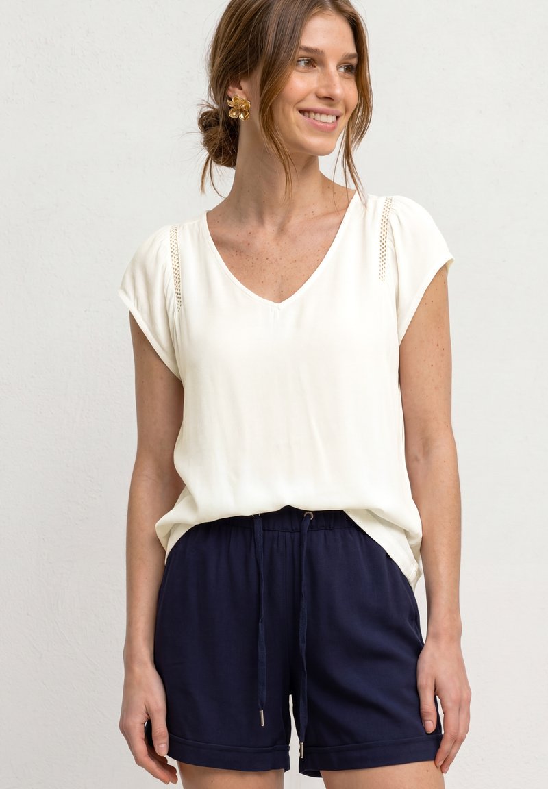 Femme aux cheveux châtain clair noués en chignon, portant des boucles d'oreilles en forme de fleurs, un chemisier blanc à manches courtes et un short bleu marine à cordon, souriante et regardant vers la droite.