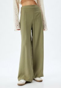 Pantalons à jambes larges en vert olive, fabriqués à partir d'un tissu doux. Présentent une taille haute et une texture légère. Associés à des baskets blanches.