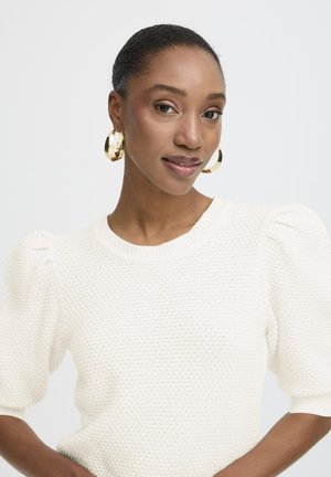 Sieviete ar gludi atpakaļ sapītu matu sakārtojumu, valkājot zelta riņķveida auskarus un baltu teksturētu īsroku džemperi, pozē pret vienkāršu fona sienu.