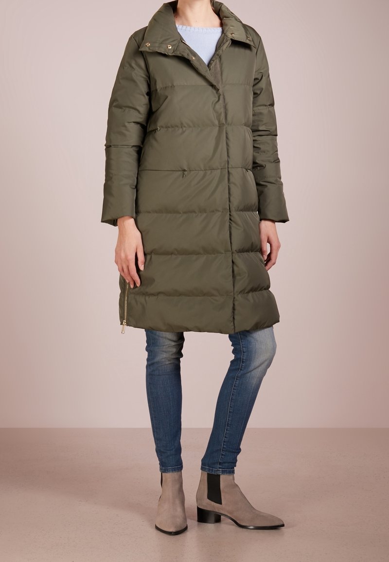 Olivgrüne Steppjacke mit hochschließendem Kragen und goldenen Hardware-Accents, ausgestattet mit langen Ärmeln und einem mittellangen Schnitt. Getragen mit blauen Jeans und grauen Stiefeletten.