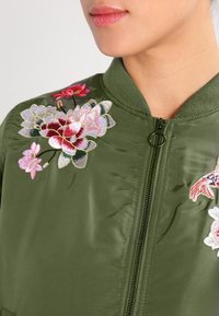 Olivově zelená bomber bunda s květinovou výšivkou v růžové a bílé. Má přední zip a žebrovaný límec a manžety.