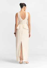 Robe maxi sans manches de couleur crème avec une taille peplum, un dos profond et une fente sur le côté, fabriquée à partir d'un tissu lisse.