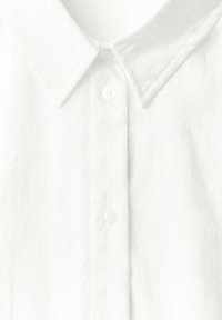 Camisa blanca de cuello con una tela texturizada, que presenta una tapeta de botones y botones lisos. La vista en primer plano resalta los detalles y la costura.