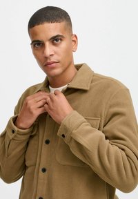 Tan fleece overhemdjack met zwarte knopen, kraag en borstzak. Model past de kraag aan, waarbij hij een gestructureerd oppervlak laat zien.