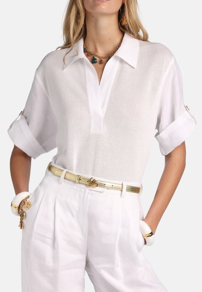 White short-sleeve polo shirt with a textured fabric, featuring a V-neck, rolled cuffs, and paired with high-waisted white trousers and a metallic belt.