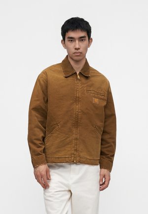 Jeune homme portant une veste marron à fermeture éclair avec col et un pantalon blanc, debout devant un fond clair uni.