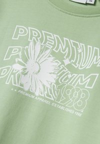 Žalias džemperis, turintis baltą grafiką su užrašu "PREMIUM" ir gėlių iliustracija. Pagamintas iš medvilnės, turinčios švelnų paviršių.