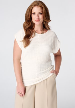 Vrouw met lang golvend rood haar, gekleed in een getextureerde witte top met korte mouwen en beige wijde broekspijpen, staand met één hand in de zak.