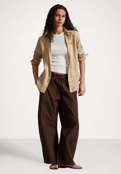 Kobieta o kręconych włosach, ubrana w podwiniętą beżową koszulę na białym topie, luźne brązowe spodnie i sandały, stojąca na białym tle.