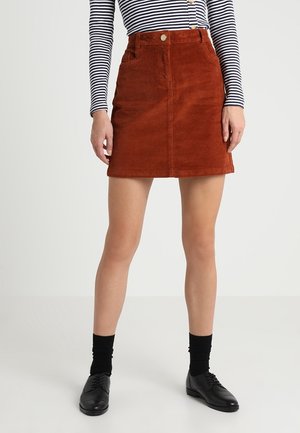 Persoon die een roestkleurige corduroy minirok, zwarte sokken, zwarte veterschoenen en een zwart-wit gestreept longsleeve met knopen draagt.