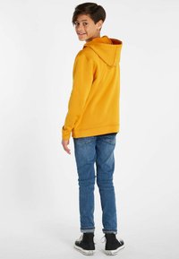 Felpa gialla con cappuccio, materiale morbido, vestibilità comoda, tasca a marsupio. Indossata con jeans blu e sneakers alte nere. Vista posteriore.