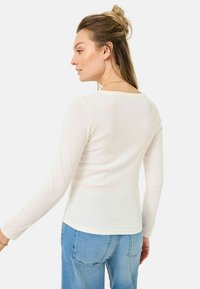 Top bianco a maniche lunghe in una texture a coste con scollatura a barchetta, abbinato a jeans in denim blu. Il tessuto aderisce strettamente al corpo.