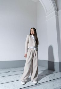 Jasno beżowa skórzana marynarka o kroju cropped i szerokie spodnie, nałożone na białą koszulę. Gładki materiał, dopasowany krój, zestawione z masywnymi białymi sneakersami.