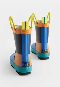 Bottes en caoutchouc colorées avec des sections orange, bleue et noire, des poignées vertes et une étiquette "HEY!" au dos. Semelle en caoutchouc texturée.