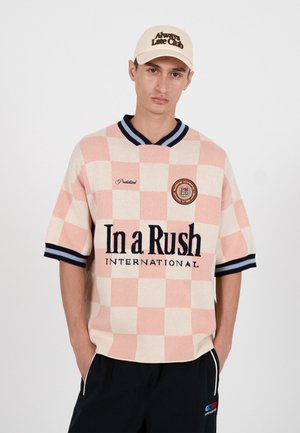 Ung mann iført beige og rosa rutete kortermet skjorte med teksten "In a Rush International" og beige caps med teksten "Always Late Club".